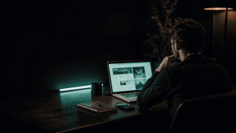 Mężczyzna pracuje przy laptopie na ciemnym biurku oświetlonym turkusowym światłem i ciepłą lampką.