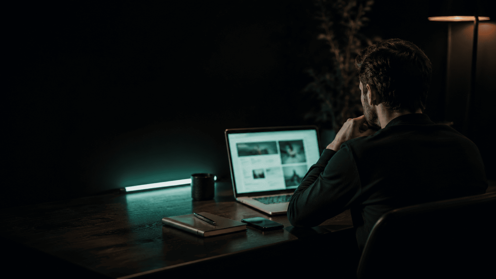 Mężczyzna pracuje przy laptopie na ciemnym biurku oświetlonym turkusowym światłem i ciepłą lampką.
